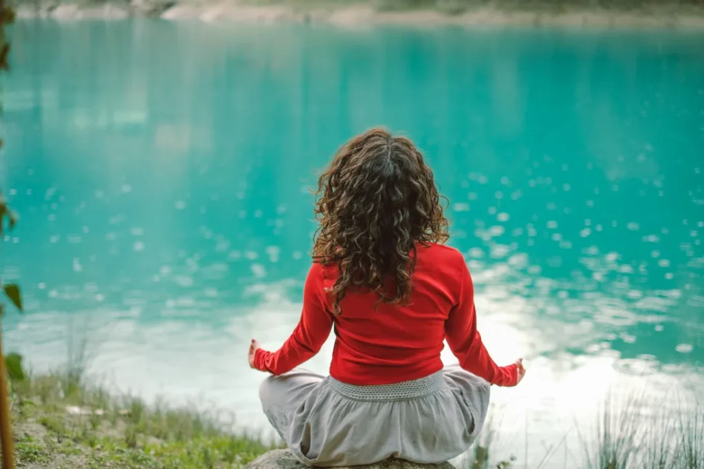 Woman seated on a rock by the water, meditating with her back to the camera